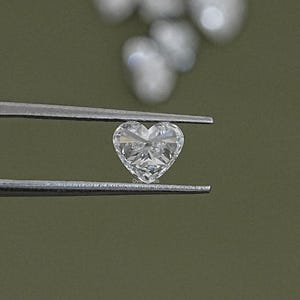 May include: A clear heart-shaped diamond held by tweezers against a green background.