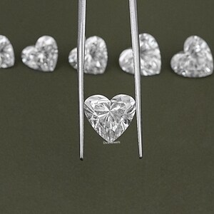 May include: A close-up of a heart-shaped diamond held by tweezers. The diamond is clear and sparkly, and it is surrounded by other heart-shaped diamonds.