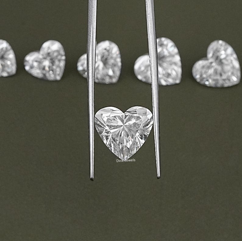 May include: A close-up of a heart-shaped diamond held by tweezers. The diamond is clear and sparkly, and it is surrounded by other heart-shaped diamonds.