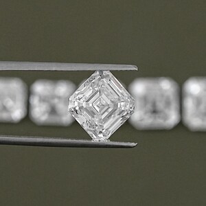 May include: A clear, square-shaped diamond with a step-cut, held by tweezers. The diamond is surrounded by other smaller diamonds.