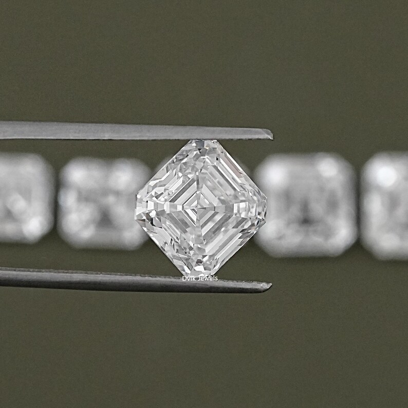 May include: A clear, square-shaped diamond with a step-cut, held by tweezers. The diamond is surrounded by other smaller diamonds.
