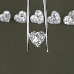 May include: A clear heart-shaped diamond held by tweezers against a green background. The diamond is faceted and reflects light.