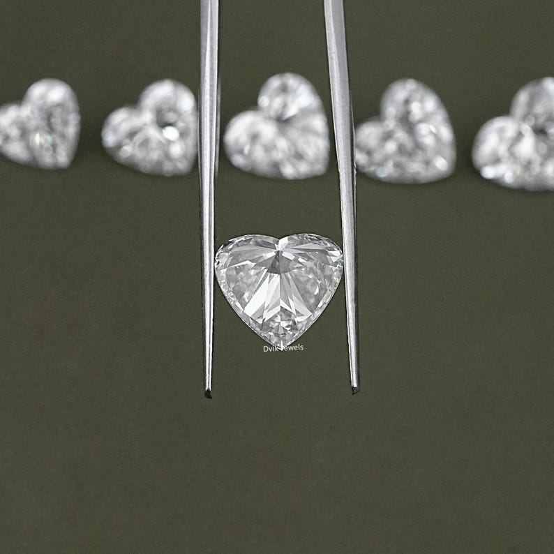 May include: A clear heart-shaped diamond held by tweezers against a green background. The diamond is faceted and reflects light.