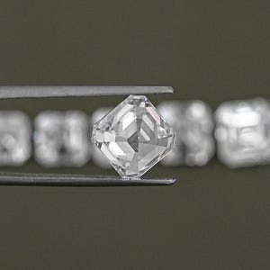 May include: A clear, square-shaped diamond gemstone held in tweezers. The diamond is faceted and has a brilliant cut.