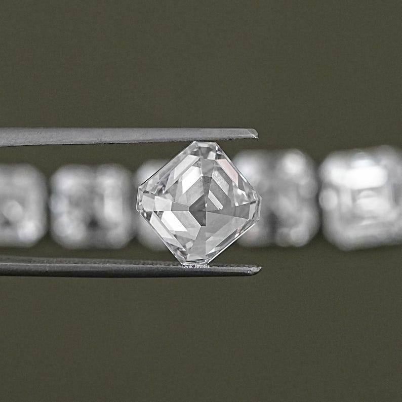 May include: A clear, square-shaped diamond gemstone held in tweezers. The diamond is faceted and has a brilliant cut.