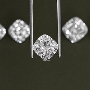 K&ouml;nnte beinhalten: Ein klarer, quadratischer Diamant, der mit einer Pinzette vor einem dunkelgr&uuml;nen Hintergrund gehalten wird. Der Diamant hat einen Kissen-Schliff und ist von kleineren Diamanten umgeben. Der Text "Dvik Jewels" ist unter dem Diamanten sichtbar.