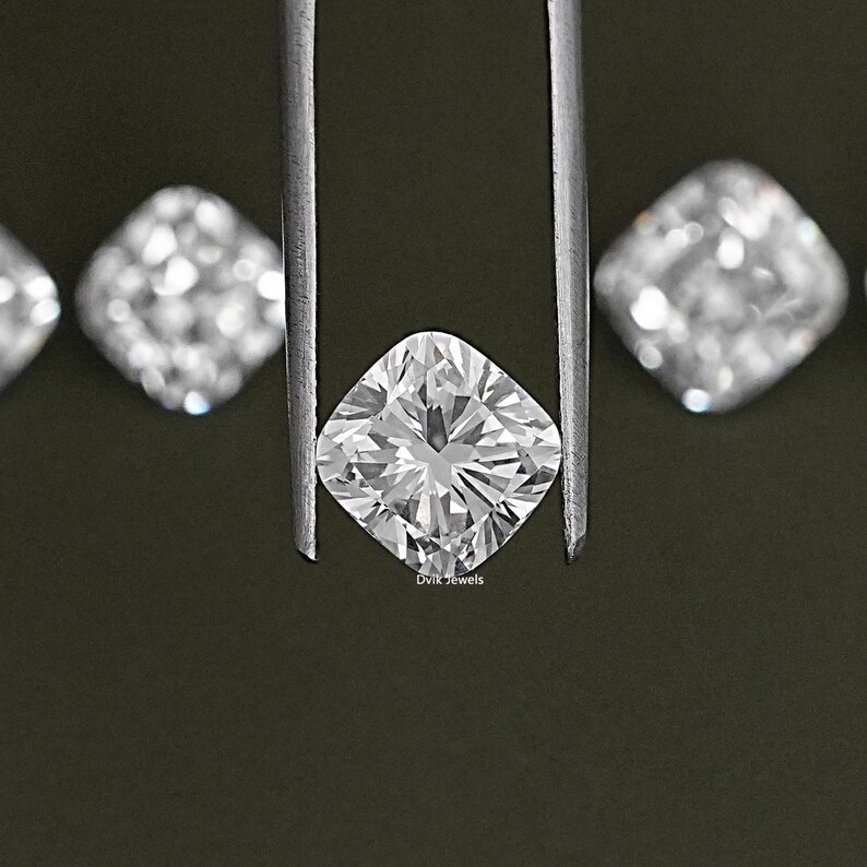 K&ouml;nnte beinhalten: Ein klarer, quadratischer Diamant, der mit einer Pinzette vor einem dunkelgr&uuml;nen Hintergrund gehalten wird. Der Diamant hat einen Kissen-Schliff und ist von kleineren Diamanten umgeben. Der Text "Dvik Jewels" ist unter dem Diamanten sichtbar.