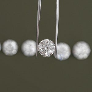 May include: A round, clear diamond held by tweezers. The diamond is surrounded by smaller, round diamonds.