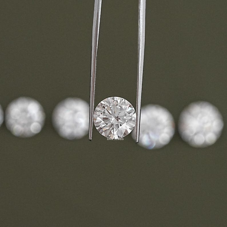 May include: A round, clear diamond held by tweezers. The diamond is surrounded by smaller, round diamonds.