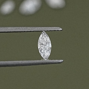 May include: A clear, marquise-shaped diamond held by tweezers against a green background.