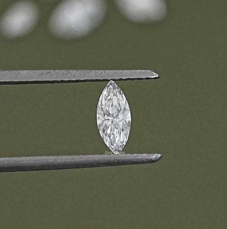 May include: A clear, marquise-shaped diamond held by tweezers against a green background.