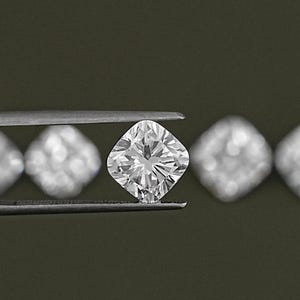 K&ouml;nnte beinhalten: Ein klarer, quadratischer Diamant wird in einer Pinzette gehalten. Der Diamant ist von anderen kleineren, unscharfen Diamanten umgeben.