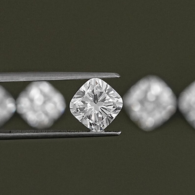 K&ouml;nnte beinhalten: Ein klarer, quadratischer Diamant wird in einer Pinzette gehalten. Der Diamant ist von anderen kleineren, unscharfen Diamanten umgeben.