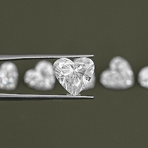 May include: A close-up of a clear heart-shaped diamond held by tweezers. The diamond is surrounded by other heart-shaped diamonds in the background.