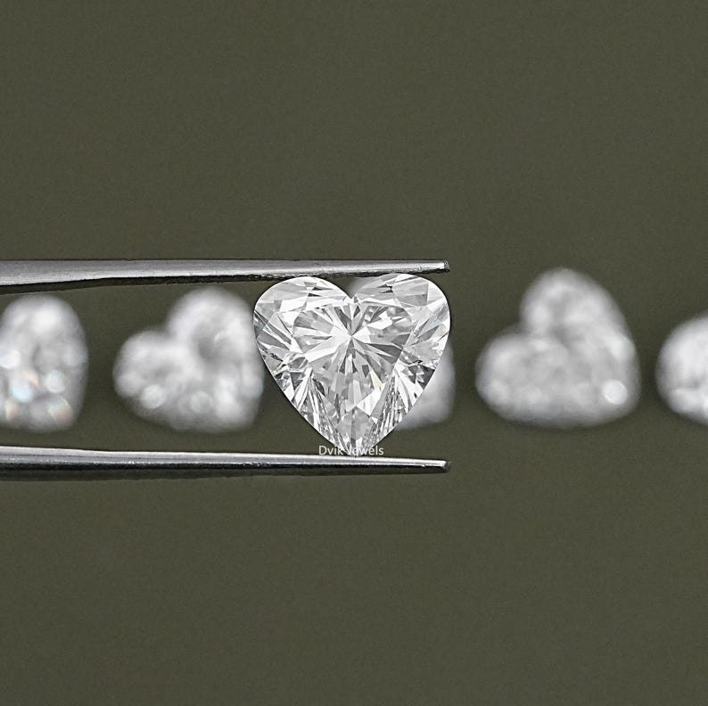 May include: A close-up of a clear heart-shaped diamond held by tweezers. The diamond is surrounded by other heart-shaped diamonds in the background.