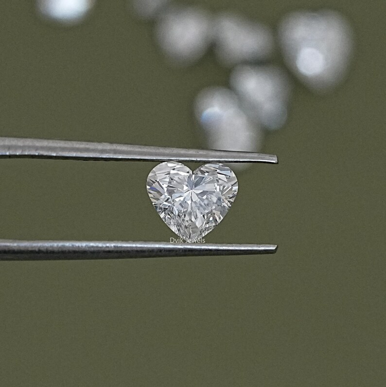May include: A heart-shaped diamond gemstone held by tweezers. The diamond is clear and sparkly, with many facets.