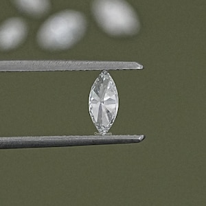 May include: A clear marquise-shaped diamond held by tweezers against a green background.