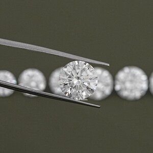 May include: A close-up of a round, clear diamond held in tweezers. The diamond is surrounded by other diamonds in the background.