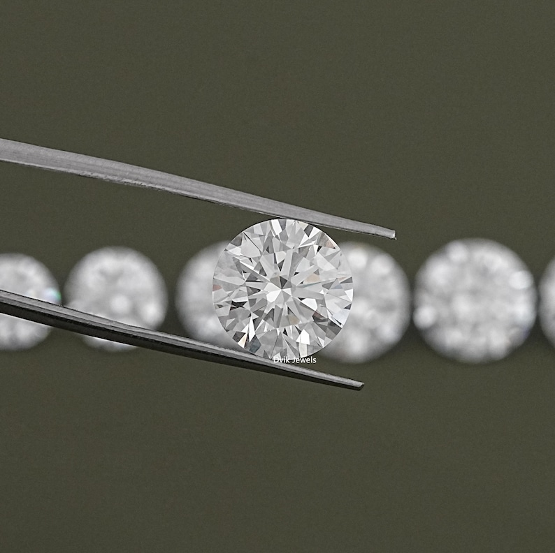 May include: A close-up of a round, clear diamond held in tweezers. The diamond is surrounded by other diamonds in the background.