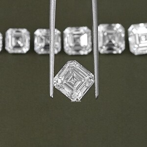 May include: A clear, square-shaped diamond gemstone held by tweezers. The diamond has a faceted cut and is surrounded by other similar diamonds.