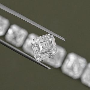 May include: A clear, square-shaped diamond gemstone with a step-cut facet pattern. The diamond is held by tweezers and is surrounded by a row of similar diamonds.