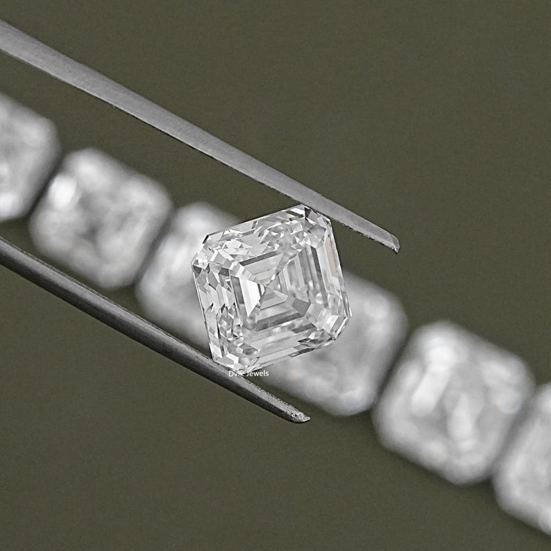 May include: A clear, square-shaped diamond gemstone with a step-cut facet pattern. The diamond is held by tweezers and is surrounded by a row of similar diamonds.