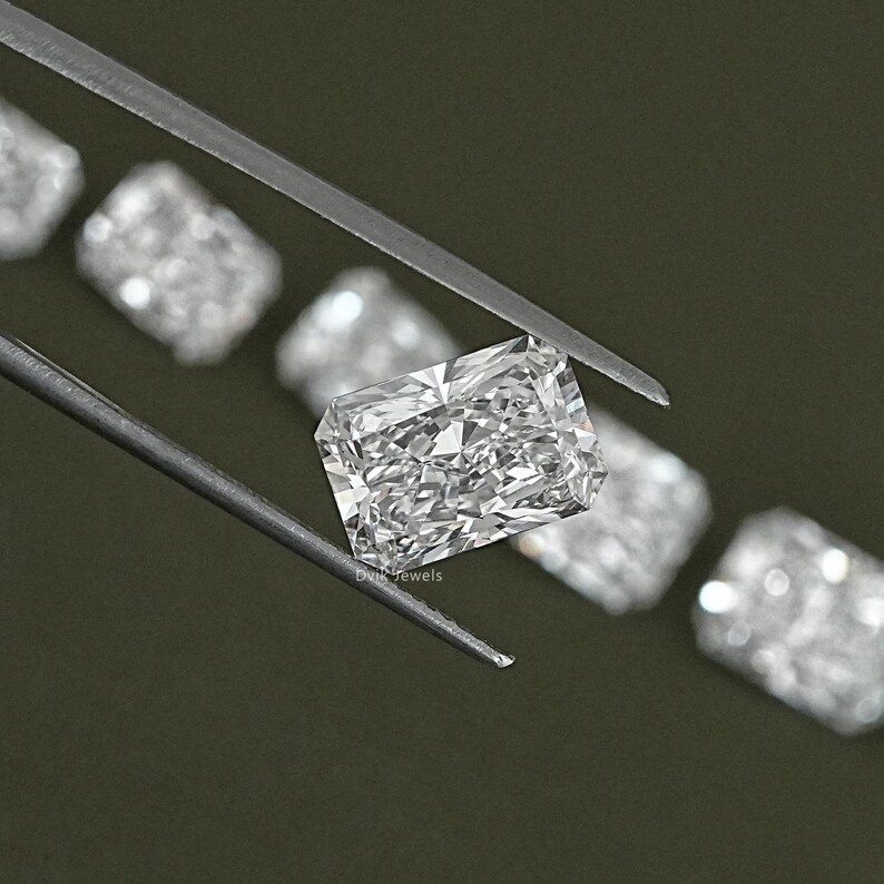 May include: A close-up of a clear, rectangular, faceted diamond gemstone. The diamond is held by a pair of tweezers and is surrounded by other diamonds.
