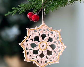 Puede incluir: Adorno navideño de copo de nieve tejido a ganchillo en rosa claro, bordeado con hilo dorado, colgado de una rama de pino con bayas rojas. Decoración navideña festiva.