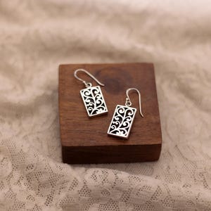 May include: A pair of silver earrings with a rectangular design featuring a delicate, intricate floral pattern.