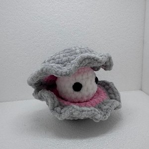May include: A crocheted oyster shell in grey with a pink interior, holding a white pearl with black eyes. The plush shell has a textured appearance, creating a soft, tactile aesthetic. A whimsical, handmade item.