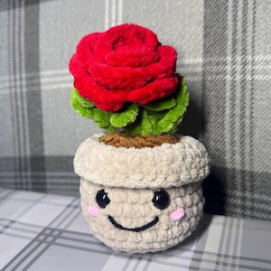 May include: A crocheted plant featuring a red rose and green leaves in a beige pot with a smiling face. The pot has pink cheeks and black eyes. The rose is made from soft yarn, creating a plush texture.