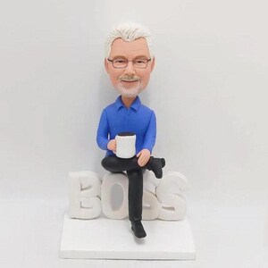 Könnte beinhalten: Eine benutzerdefinierte Figur eines Menschen, der auf dem Wort "BOSS" sitzt und eine Tasse Kaffee in der Hand hält. Die Figur trägt ein blaues Hemd und eine schwarze Hose.