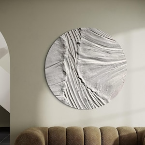 May include: A brown, tufted sofa with a large, round, white wall art piece featuring a textured, abstract design. The sofa is in front of a white wall and a black and white patterned rug.