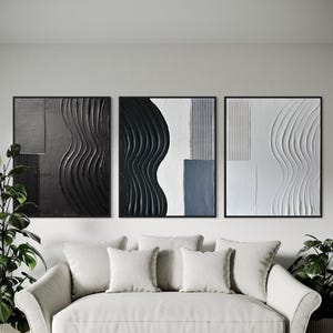 Puede incluir: Tres obras de arte murales abstractas enmarcadas. La obra presenta diseños monocromáticos texturizados en negro, blanco y gris. Cada pieza tiene un marco negro y un patrón único de formas curvas y geométricas. El arte se exhibe sobre un sofá blanco.