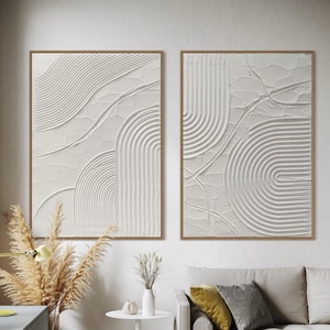 Könnte beinhalten: Zwei gerahmte abstrakte Wandkunstwerke. Jedes hat eine strukturierte, cremefarbene Oberfläche mit geschwungenen und linearen Mustern. Die Rahmen sind warmbraun, und das Kunstwerk wird in einem hellen, modernen Wohnraum ausgestellt.