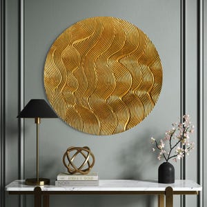 Puede incluir: Una decoración de pared circular dorada con un patrón ondulado texturizado. La decoración está colgada en una pared gris sobre una mesa consola de mármol blanco con un marco de metal dorado. Una esfera de metal dorado se encuentra encima de dos libros en la mesa. Un jarrón negro con un arreglo de flores rosas está en la mesa.
