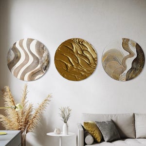 Op de afbeelding: Drie ronde wandkunstwerken met textuurontwerpen. Eén heeft golvende lijnen in beige en bruin, een andere is effen goudkleurig en de derde heeft gouden en witte patronen. De kunst wordt boven een bank tentoongesteld en voegt een moderne touch toe aan de kamer.