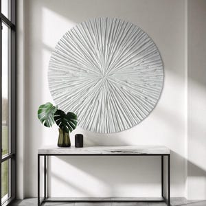 Könnte beinhalten: Ein großes, kreisförmiges, weißes Wandkunstobjekt mit einem strukturierten Strahlenmuster, bei dem Linien von der Mitte ausgehen. Das Kunstwerk ist an einer schlichten Wand über einem rechteckigen Konsolentisch mit weißer Marmorplatte angebracht.
