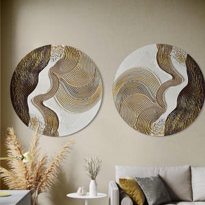 Op de afbeelding: Twee ronde wandkunstwerken. Elk heeft een wit midden met goud- en beige concentrische lijnen. Donkerbruine en goudkleurige getextureerde secties omlijsten het witte midden. De kunst heeft een modern, abstract ontwerp.