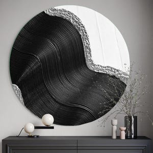 Op de afbeelding: Een groot, rond kunstwerk voor aan de muur met een opvallend ontwerp. Het kunstwerk heeft een zwart, golfachtig patroon met zilveren accenten langs de rand. Het stuk is aan een muur bevestigd boven een donkergrijze kast, met decoratieve items.