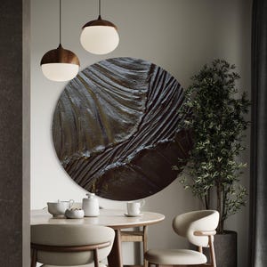 Op de afbeelding: Een groot, rond, getextureerd kunstwerk aan de muur met een donker, aards kleurenpalet domineert de scène. Het kunstwerk hangt boven een ronde eettafel met stoelen in neutrale tinten. Twee hanglampen met houten toppen en witte bollen hangen boven de tafel.