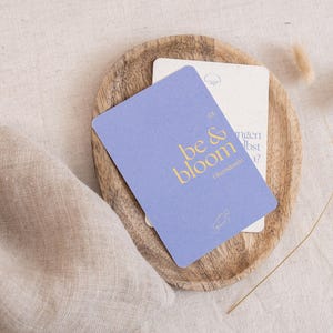 Könnte beinhalten: Eine hellviolette Karte mit den Worten "be & bloom" in goldfarbenen Buchstaben liegt auf einem Holztablett, mit einer zweiten Karte dahinter. Das Tablett liegt auf einer beigefarbenen Leinenoberfläche, mit getrockneten Blumen und einem Leinentuch.