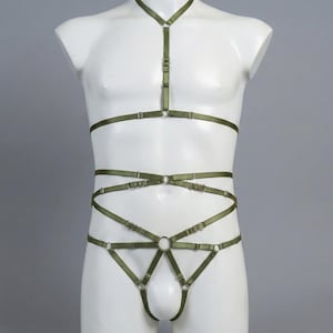 May include: An olive green body harness and thong set on a white mannequin. The harness has adjustable straps and metal ring details. The thong has a triangular design with a ring detail. The set is made of a smooth, satin-like material.