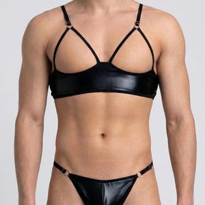 May include: Black lingerie set featuring a bra with thin straps and a matching thong. The bra has a unique design with open cups and a shiny, faux leather appearance. The thong has a similar shiny finish and ring details.