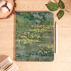 May include: A rectangular notebook with a Monet-style water lilies painting in green, blue, and pink. A white stylus lies beside the notebook on a light wood surface. A small stack of white notepaper and a wood slice with cotton are also visible.