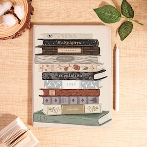 May include: A cream-colored notebook featuring a stack of illustrated books. The book titles include "Midnights," "Evermore," "Folklore," "Lover," "Reputation," "1989," "Red," "Speak Now," and "Fearless." A white stylus rests beside the notebook.