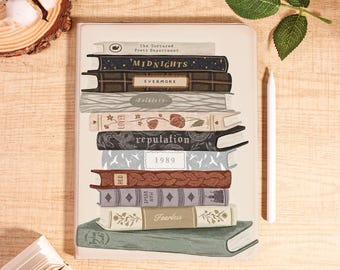 May include: A cream-colored notebook featuring a stack of illustrated books. The book titles include "Midnights," "Evermore," "Folklore," "Lover," "Reputation," "1989," "Red," "Speak Now," and "Fearless." A white stylus rests beside the notebook.