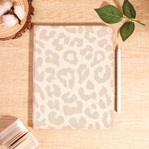 Può includere: Una custodia beige per tablet con un motivo leopardato grigio chiaro. Una penna bianca è appoggiata accanto alla custodia su una superficie di legno chiaro. Elementi decorativi includono cotone e una foglia verde.