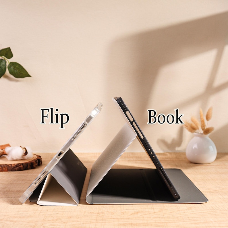 May include: Two tablet cases, one silver and one black, are displayed open on a wooden surface. The silver case is labeled "Flip," and the black case is labeled "Book." A small white vase with dried flowers is in the background.