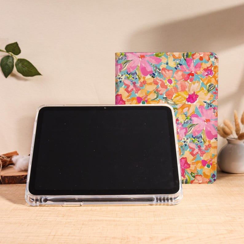 May include: A clear tablet case with a black screen, a floral-patterned journal, and a small vase with dried flowers. The journal features a colorful design with pink, yellow, and blue flowers. The items are on a wooden surface.